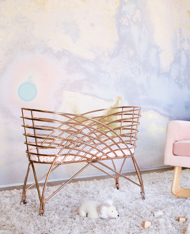 A gold-colored, modern crib on a fluffy white rug, with a toy mouse nearby. The background features a pastel abstract wall design, and part of a pink armchair is visible on the right.
